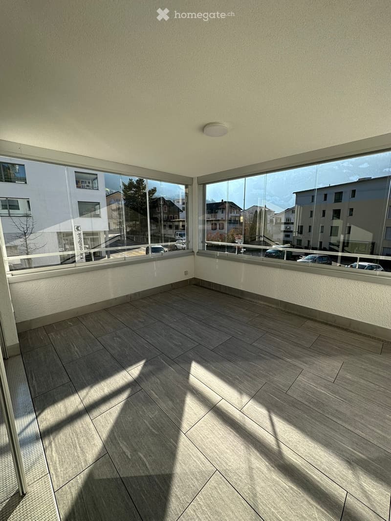 Empty balcony with wooden flooring, large glass windows, and a view of the street and surrounding buildings.