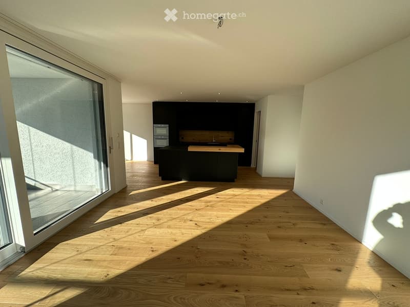 Bright, open-plan space with light wooden flooring, large glass sliding doors leading to a balcony, and a modern kitchen area with dark cabinetry and a wooden countertop.