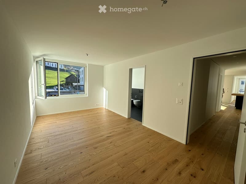 Empty room with light wooden flooring, white walls, and a large window with an open view of the outside. A doorway leads to a bathroom with a toilet and sink visible.