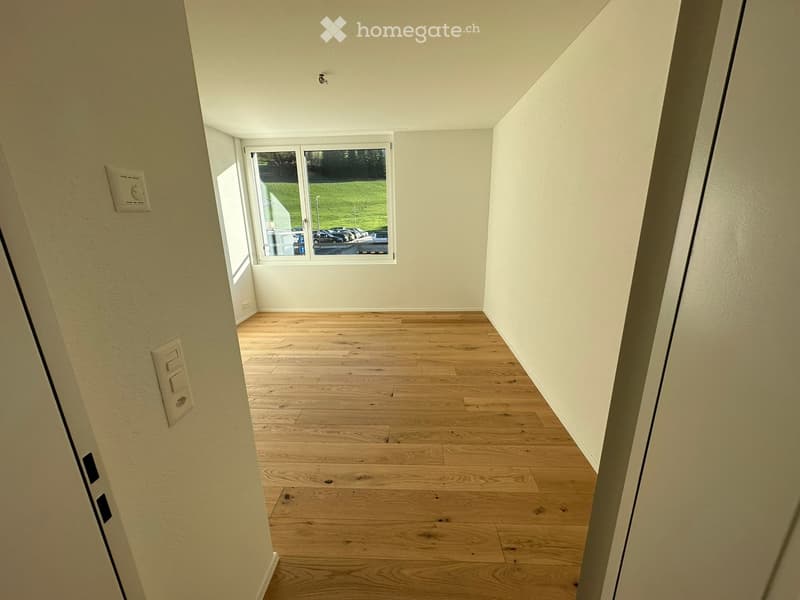 Empty room with wooden flooring and white walls, featuring a large window with a view of a green area and parked cars.