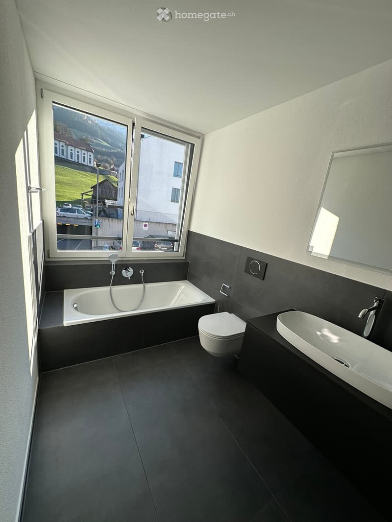 Modern bathroom with dark grey tiles, a bathtub, toilet, sink, and large windows offering a view of the outdoor area.