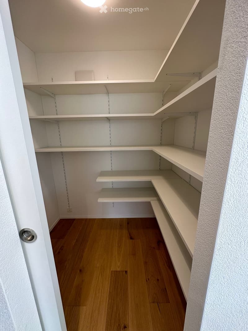 Empty closet with white shelves and wooden flooring, featuring built-in storage compartments and a closed door.