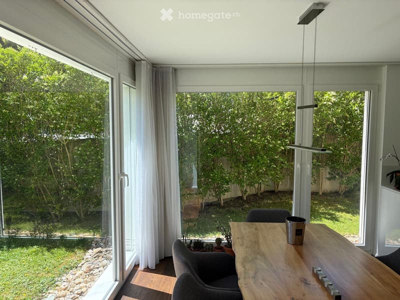 Bright room with large windows and a view of a garden, featuring a wooden table, chairs, and potted plants.
