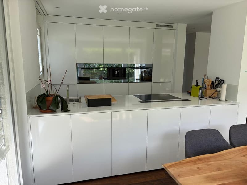 Modern kitchen with white cabinets, a central island, and a wooden dining table with chairs. Features include a sink, faucet, cutting board, and various kitchen utensils.