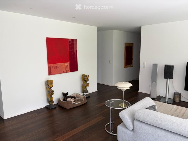 Modern living room with hardwood flooring, white walls, grey sofa, glass coffee table, floor lamp, dog bed, and decorative sculptures.