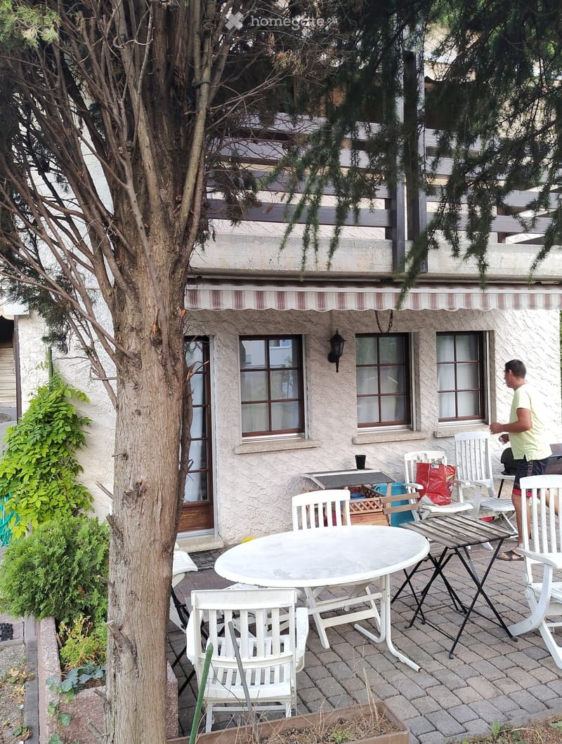 white stucco exterior, brown wood awning, large windows, balcony, front garden, patio with white chairs and tables