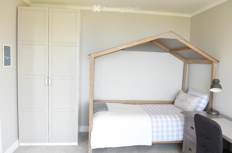 single bed with canopy, white sheet and blue checked blanket, white pillows, lamp on the desk, wardrobe with double doors, framed picture on the wall