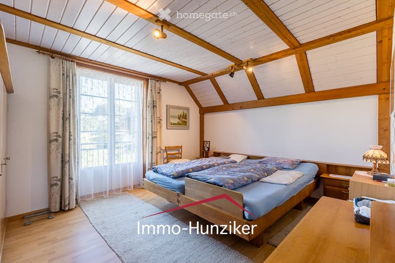 Cozy attic bedroom with wooden beams, slanted ceiling, large window with curtains, double bed with blue bedding, nightstand, chair, and a small table with a lamp.