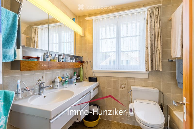 Bathroom with beige tiled walls, a double sink, a toilet, a mirror with wooden cabinet, and a window with a curtain. Personal care items are on the sink counter.