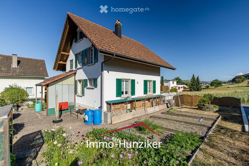 Two-story house with white walls, green shutters, and a tiled roof. The house features a garden area with raised beds, a wooden fence, and a small patio with outdoor furniture.
