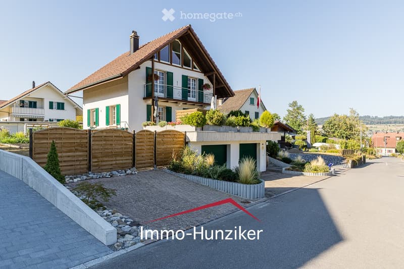 A two-story house with a red-tiled roof, white walls, green shutters, and a balcony. The house features a wooden fence, a driveway, and surrounding greenery.