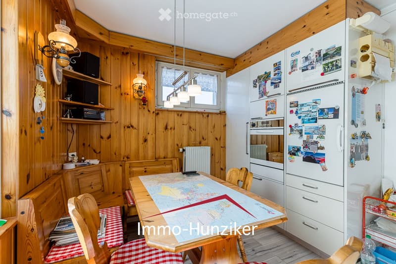 Cozy kitchen with wooden walls and ceiling, featuring a dining table with chairs, a refrigerator covered with photos, and a lamp hanging from the ceiling.