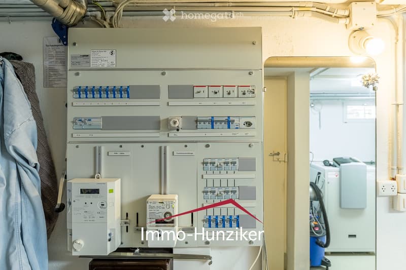 Electrical panel with multiple switches, meters, and fuses mounted on a wall. A small section of a room with a washing machine is visible in the background.