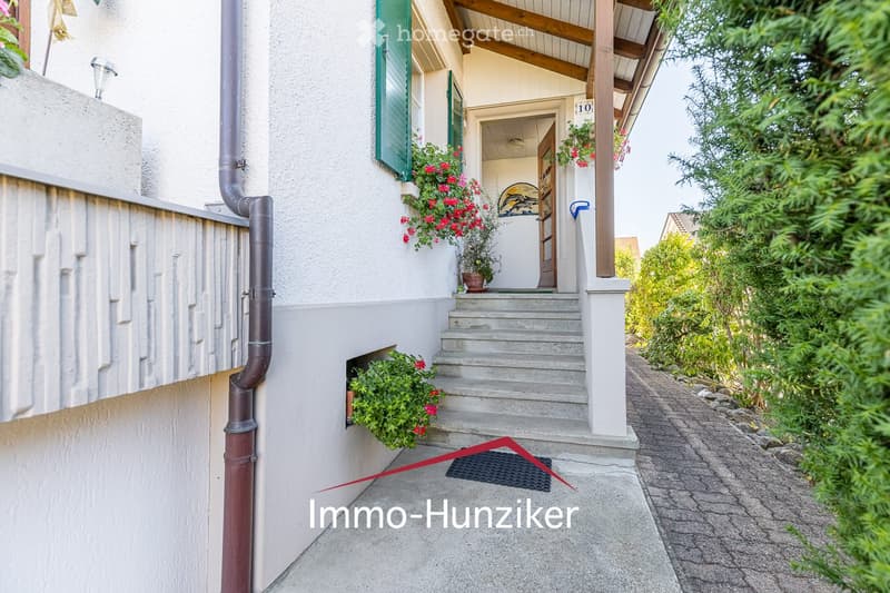 A two-story house with a raised ground floor, featuring a covered entrance with wooden beams, green shutters, and flower boxes. The pathway is paved, and there are plants and shrubs along the sides.