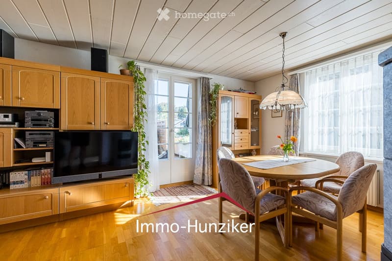 Bright living room with wooden flooring, large windows, a television on a wooden cabinet, a round dining table with chairs, and a hanging lamp.