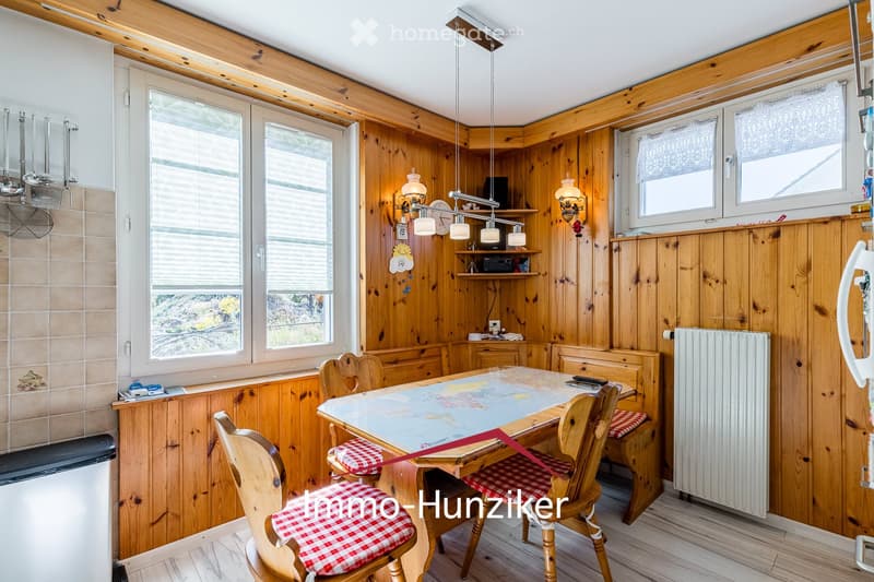 Cozy kitchen with wooden walls and ceiling, featuring a dining table with chairs, a radiator, and shelves with various items.