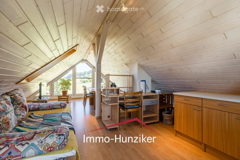 Bright, wooden-floored room with a slanted wooden ceiling, featuring a sofa, small table, kitchen area with cabinets and countertop, and a desk with chairs.