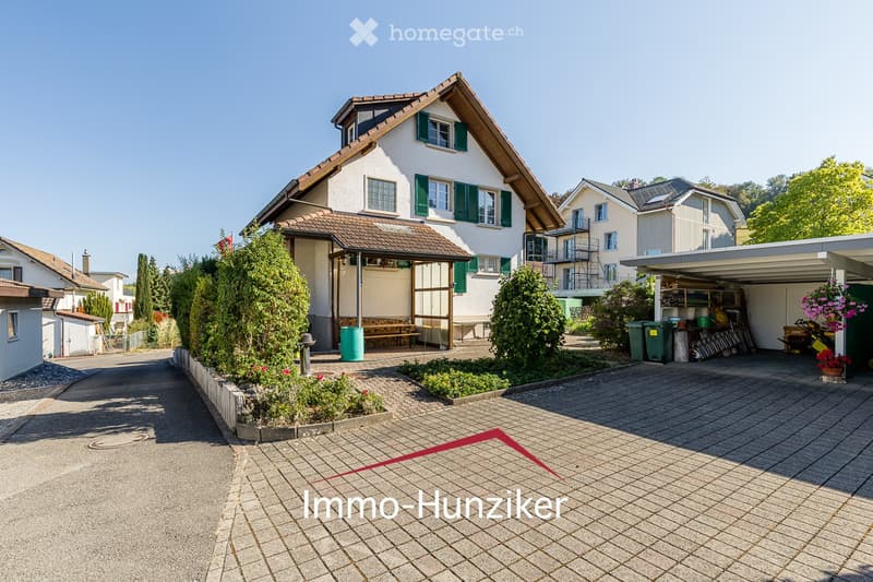 A two-story house with a tiled roof and green shutters, surrounded by a well-maintained garden and driveway. The house features a covered porch area with outdoor seating and a small garden bed in front.