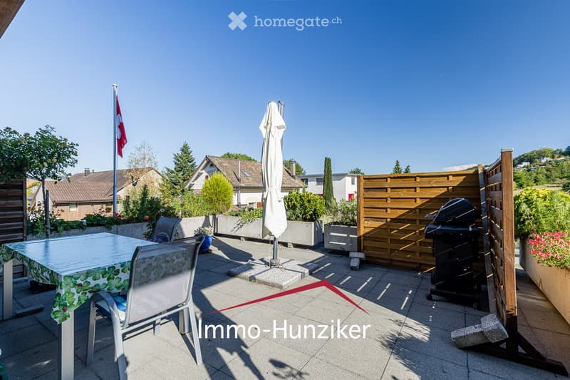 A sunny outdoor patio with a table, chairs, umbrella, and barbecue, surrounded by wooden fencing and greenery.