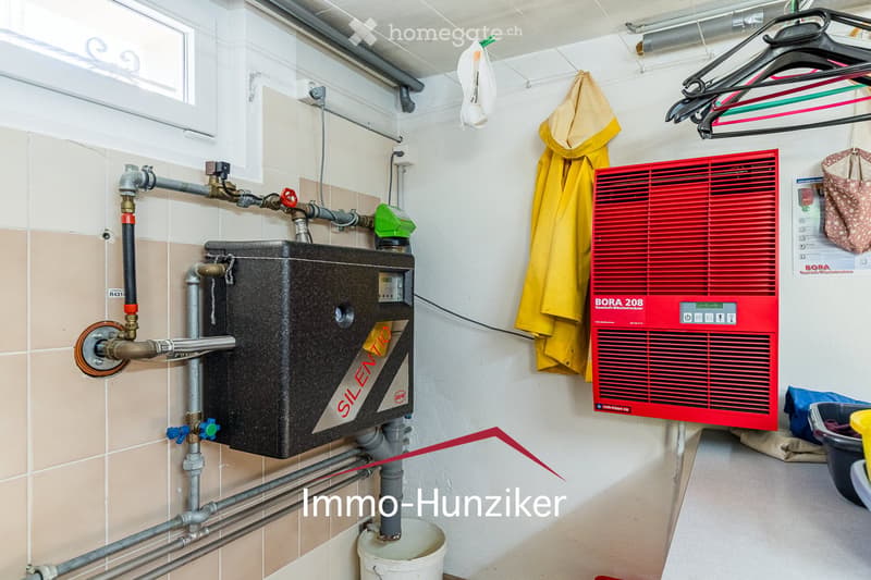 A utility bathroom with a water heater, pipes, valves, a coat hanger with a coat, and a small window.