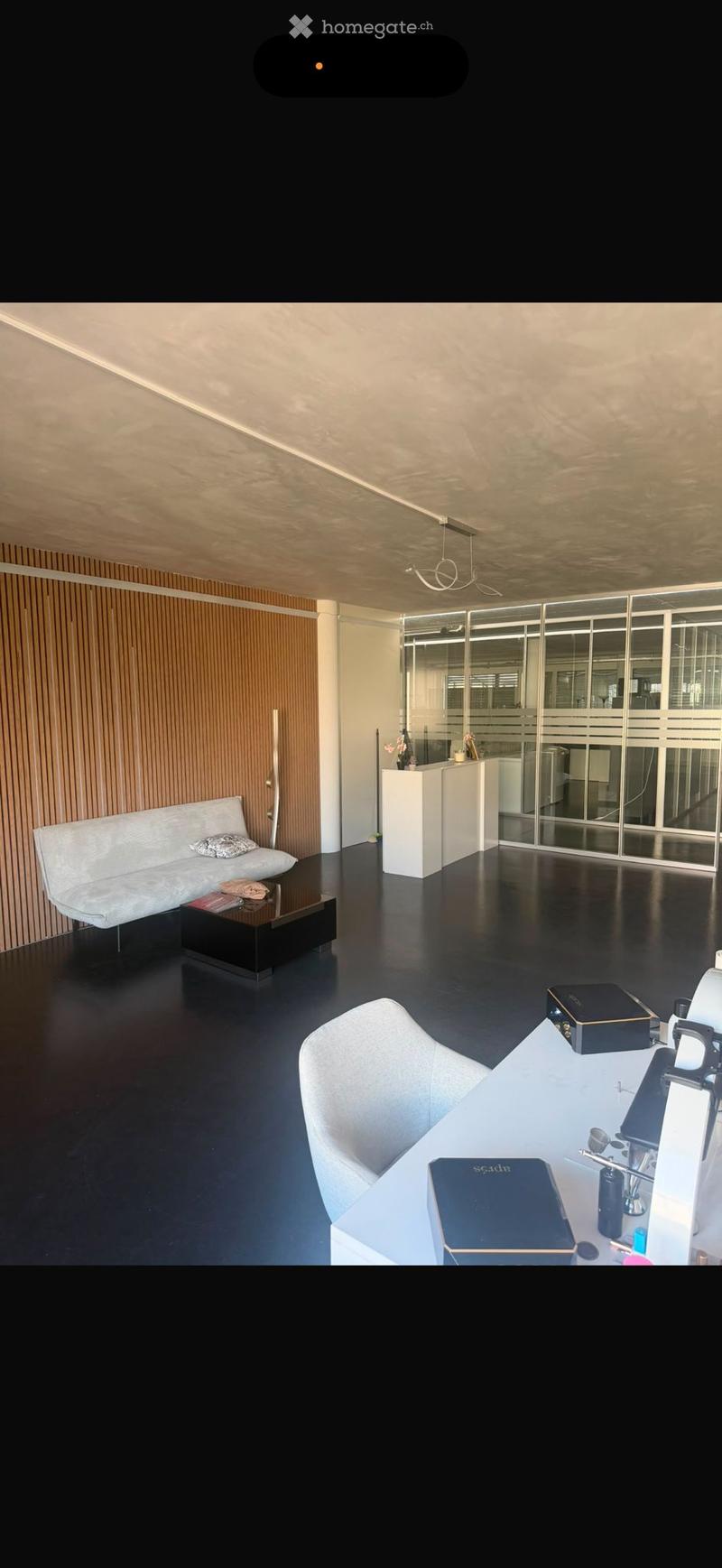 Modern, minimalist room with a white sofa, coffee table, desk, and chair. Features a sleek black floor, wooden panel wall, and glass partition.