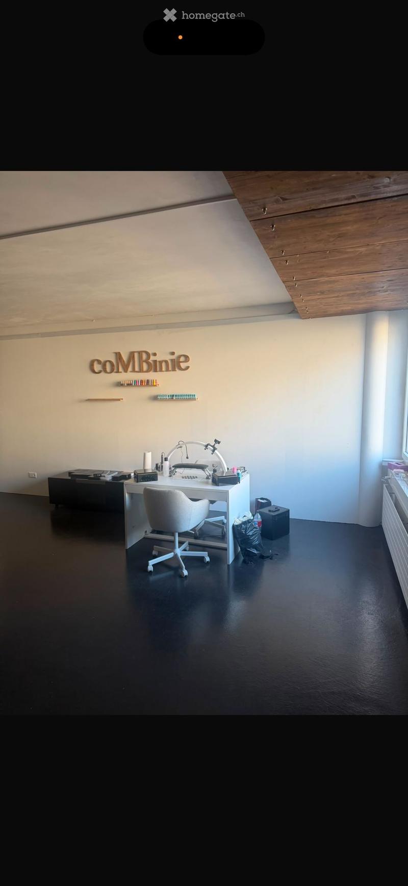 Modern, minimalist room with a white desk, white chair, and a sleek lamp. The room features a dark, polished floor and light-colored walls with a wooden slanted ceiling. Shelves with small items are mounted on the wall.