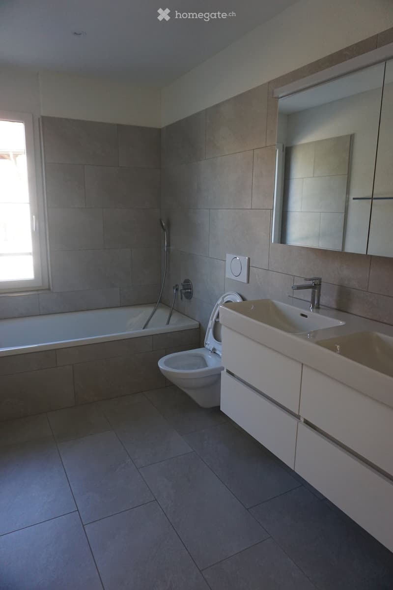 Modern bathroom with a bathtub, toilet, sink, and large mirror. Grey tiles on walls and floor, sleek white cabinetry, and a window providing natural light.