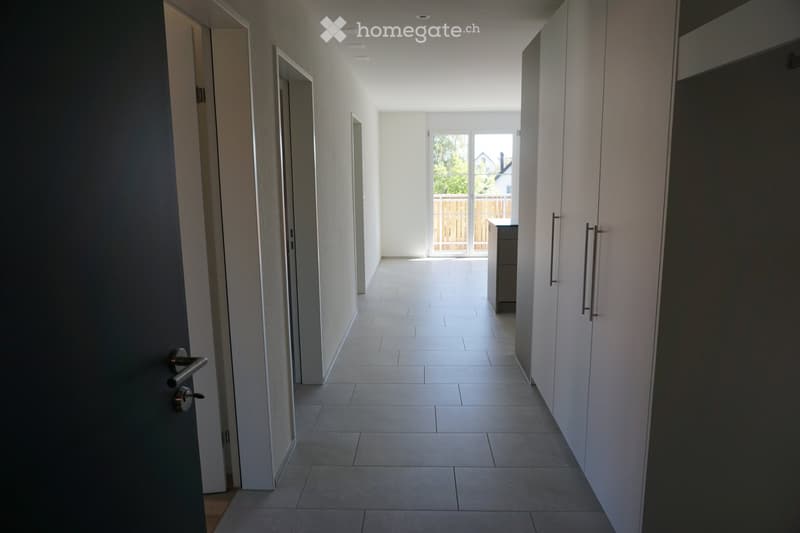 Modern hallway with tiled floor, white walls, and built-in storage units on one side. Natural light enters from the end of the corridor through a large window and sliding doors leading to an outdoor area.