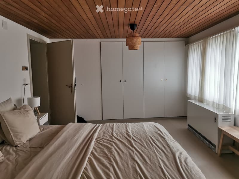 Modern bedroom with a large bed, nightstand, lamp, and built-in white wardrobe. Wooden ceiling and large window with white curtains.