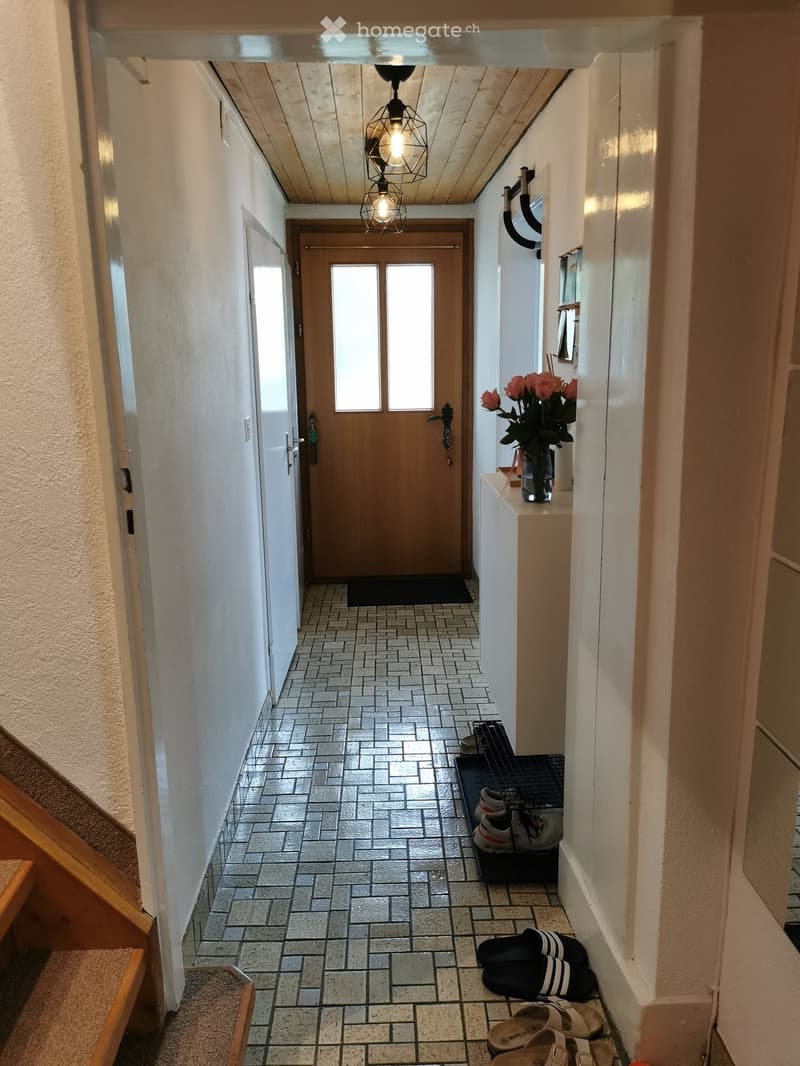 A narrow hallway with tiled flooring, white walls, and a wooden door at the end. A small console table with a vase of flowers is visible on the right side, along with a shoe rack holding several pairs of shoes and sandals.