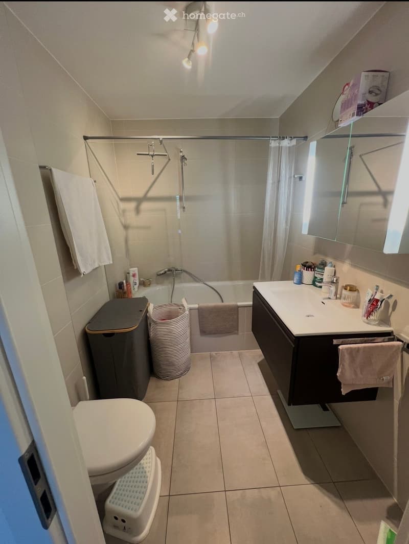 A compact bathroom with a toilet, sink, bathtub, and mirror. Features include a towel, trash bin, and a small cabinet with toiletries.