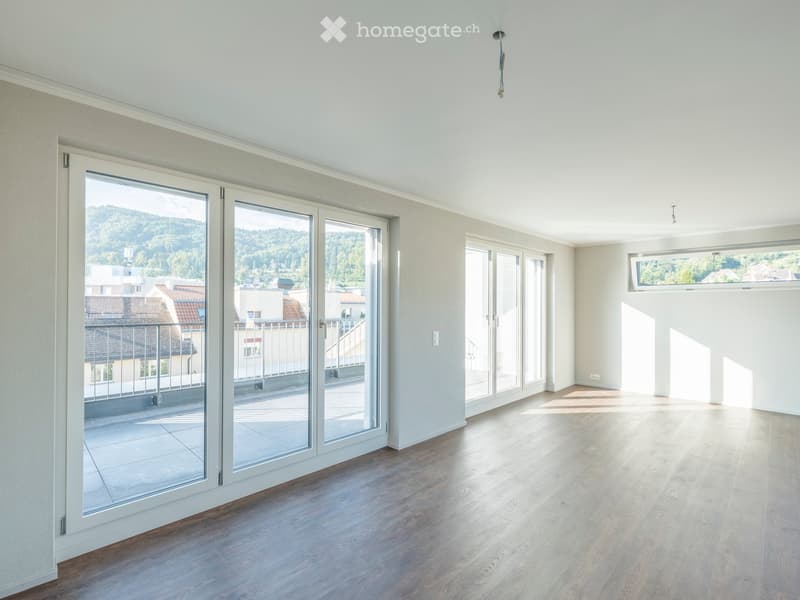 Empty room with light-colored wooden flooring, large glass sliding doors leading to a balcony, and a view of the surrounding area.