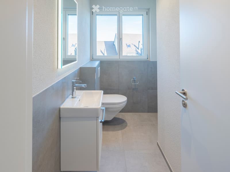 Modern bathroom with a white sink, toilet, and mirror, featuring large windows and grey tiled walls.