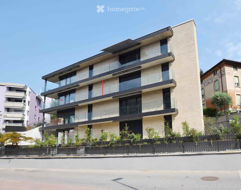 Modern multi-story building with beige facade, large windows, and balconies. Features a sleek design with glass railings and a small garden area in front.