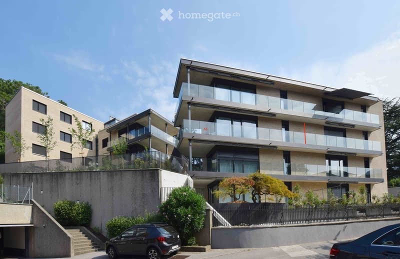Modern multi-story residential building with balconies, glass railings, and a raised ground floor entrance.