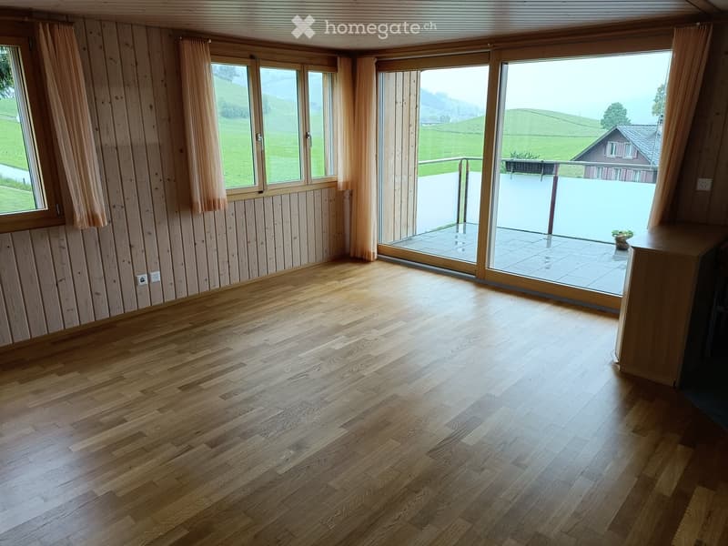 Empty room with wooden flooring, large windows with curtains, and a sliding glass door leading to a balcony.