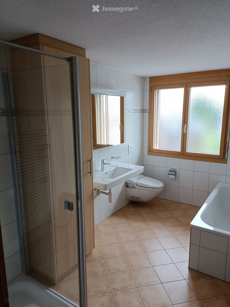 Modern bathroom with a white sink, toilet, bathtub, and large windows. Features tiled walls and wooden cabinetry.