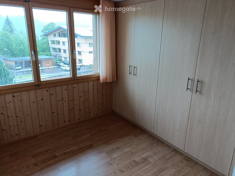Empty room with wooden flooring, light wooden walls, large windows with curtains, and built-in wardrobes.