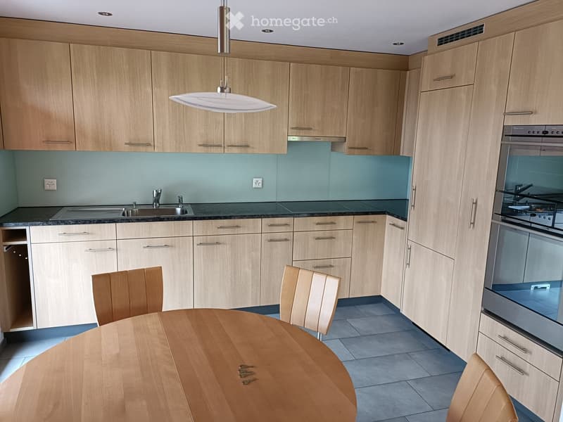 Modern kitchen with light wood cabinets, stainless steel appliances, a round wooden dining table with chairs, and a pendant light fixture.