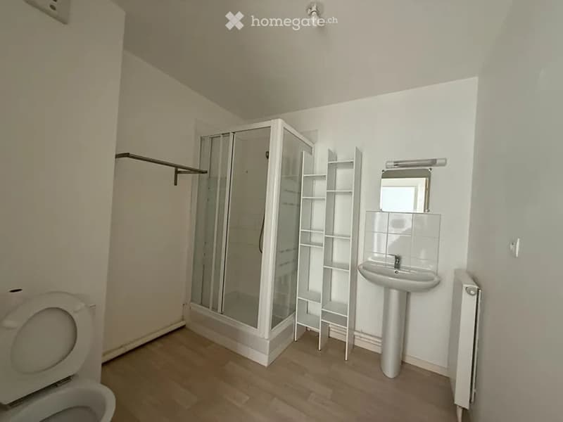 Modern bathroom with wooden flooring, white walls, a toilet, a sink with a mirror, a glass-enclosed shower, and a white shelf unit.
