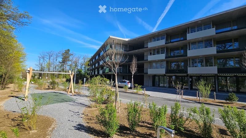 Modern multi-story residential building with balconies and large windows, surrounded by landscaped greenery and a playground area.