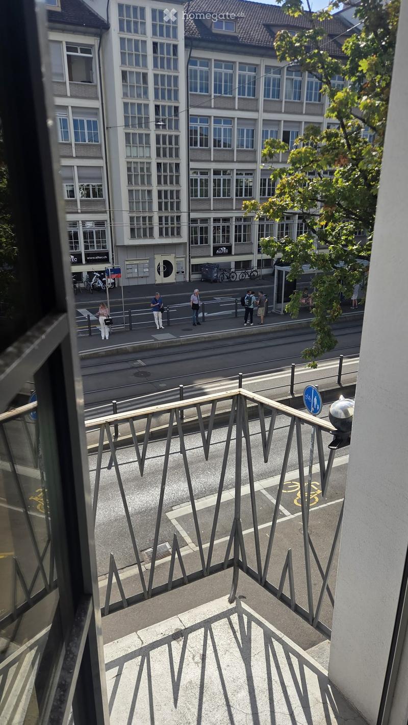 Balcony overlooking a street with modern buildings, bicycles, and pedestrians. Metal railing with geometric design.