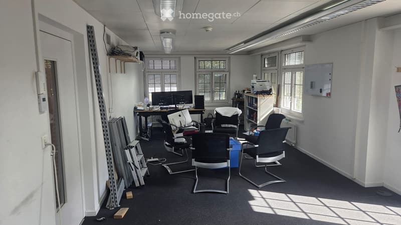 Office space with multiple chairs, a table, a screen, a printer, and a shelf. The room has large windows with blinds and is well-lit with overhead lighting.