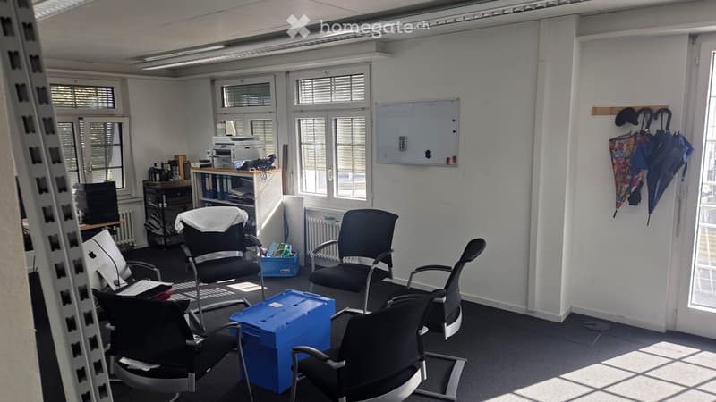 Office space with multiple black office chairs, a small table with a printer, a screen, and umbrellas hanging on the wall. The room has white walls, large windows with blinds, and a radiator.