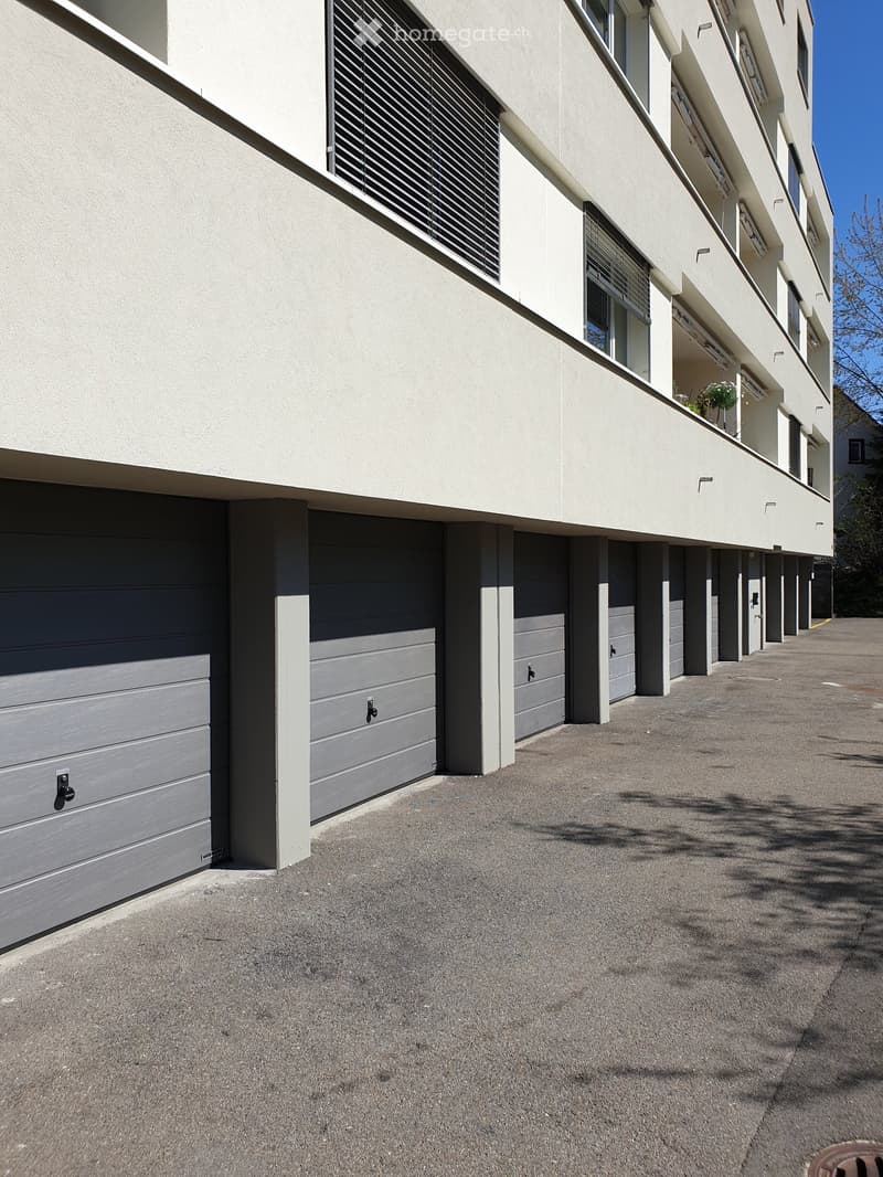 Modern multi-story building with grey garage doors and white columns, featuring windows with blinds and a small balcony with a potted plant.