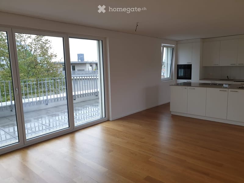 Open-plan room with light wooden flooring, white cabinets, and a built-in oven. Large sliding glass doors open to a balcony with a metal railing and tiled floor.