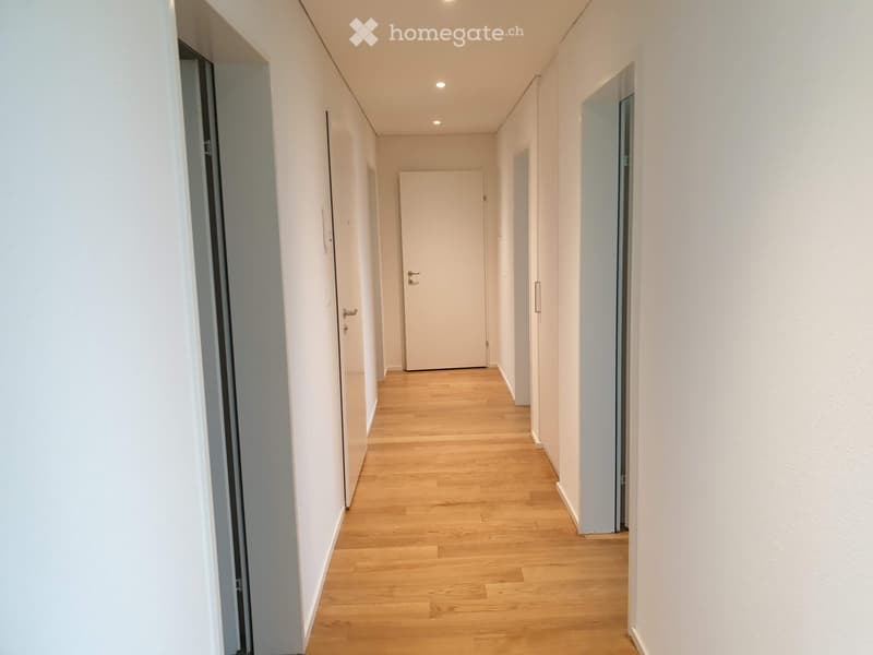 A clean, modern hallway with light wooden flooring and white walls. Features several closed doors and recessed ceiling lighting.