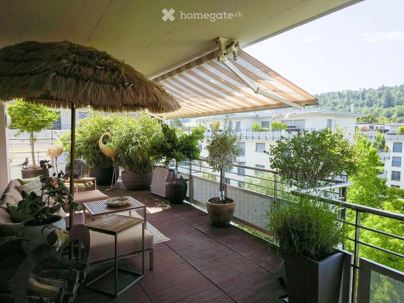 A spacious balcony with wooden decking, a large umbrella, several potted plants, a sofa, a small table, and chairs.
