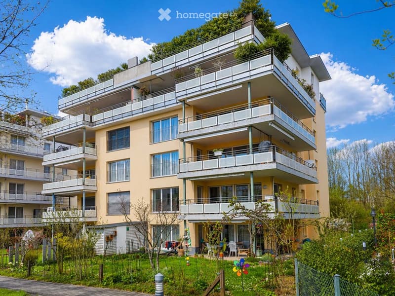 Multi-story residential building with balconies, surrounded by greenery and a garden area with outdoor furniture.