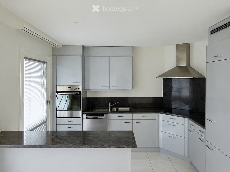 Modern kitchen with grey cabinets, stainless steel appliances, black countertops, and a large window with blinds.